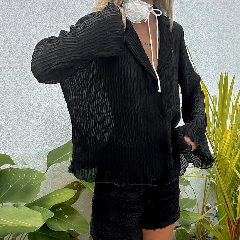 Person wearing a black textured outfit with a white lace garment underneath, standing in front of green plants.
