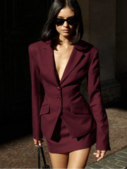 Woman wearing a burgundy suit with sunglasses, standing against a dark background.