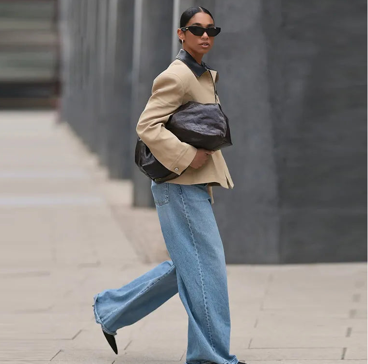 Person wearing a beige jacket, blue jeans, and sunglasses, walking on a sidewalk.