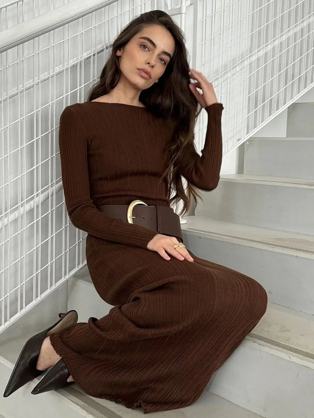 Woman in a brown ribbed dress sitting on stairs with a white wall background