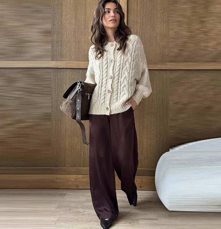 Woman wearing a cream knitted cardigan and dark pants, holding a brown leather bag against a wooden paneled wall.