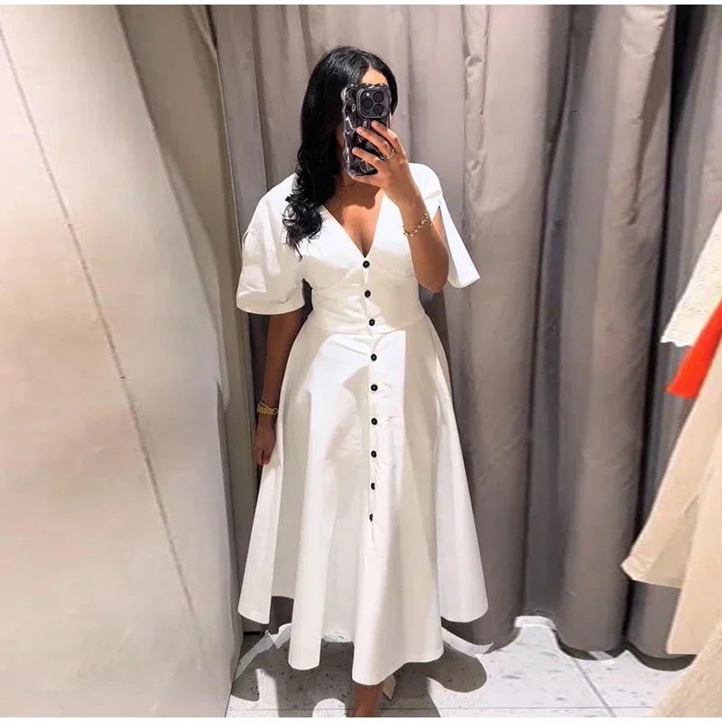 Person wearing a white dress with black buttons in front of a neutral curtain.