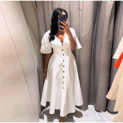 Person wearing a white dress with black buttons in front of a neutral curtain.