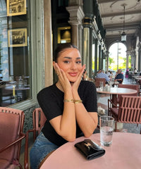 Woman sitting at a table in an outdoor cafe, posing with hands on face.