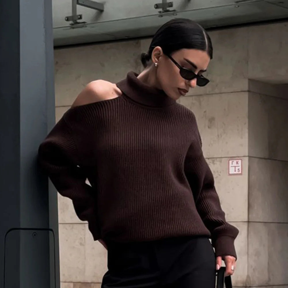 Woman wearing a brown sweater and sunglasses against a concrete wall.