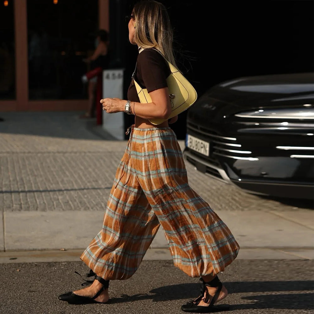 Woman walking on a street wearing plaid pants and a yellow bag.