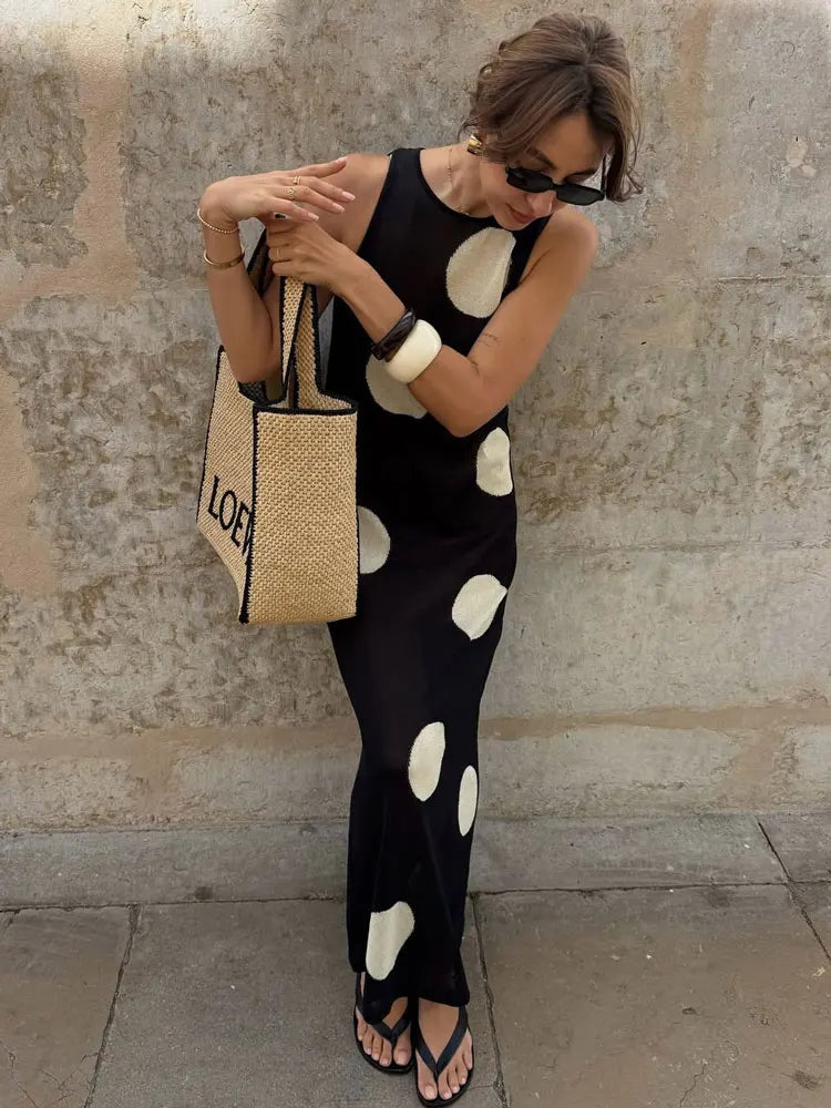 Woman in a black polka dot dress holding a straw bag against a stone wall.