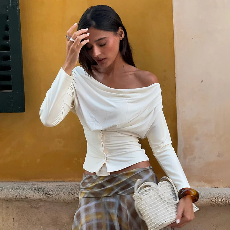 Woman in a white off-shoulder top and patterned skirt holding a woven bag against a yellow wall.