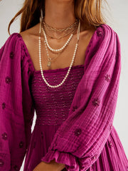 Person wearing a purple dress with a textured pattern and a matching cardigan, accessorized with necklaces.