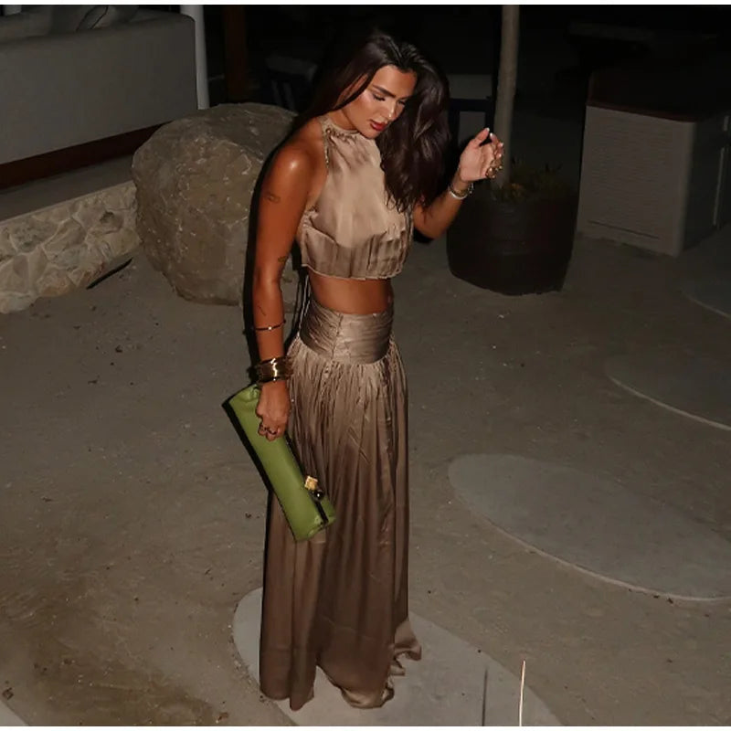 woman in a gold halter neck crop top and matching flowing maxi skirt, holding a green clutch, looking down