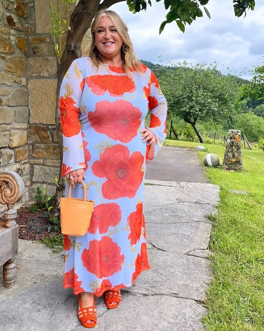 Woman in a floral dress standing outdoors with a stone wall and greenery in the background