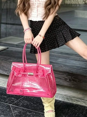 A woman holds a large, glossy pink crocodile-embossed handbag with double top handles. She is wearing a black polka-dot pleated mini skirt, a light pink top, and beige boots, standing on dark stone steps.