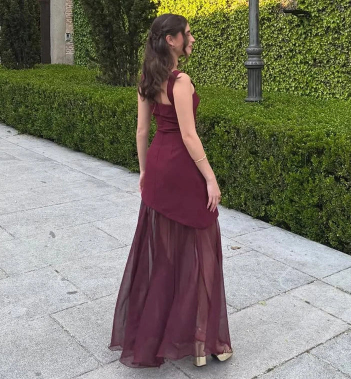 Woman in a burgundy evening gown standing on a sidewalk with greenery in the background