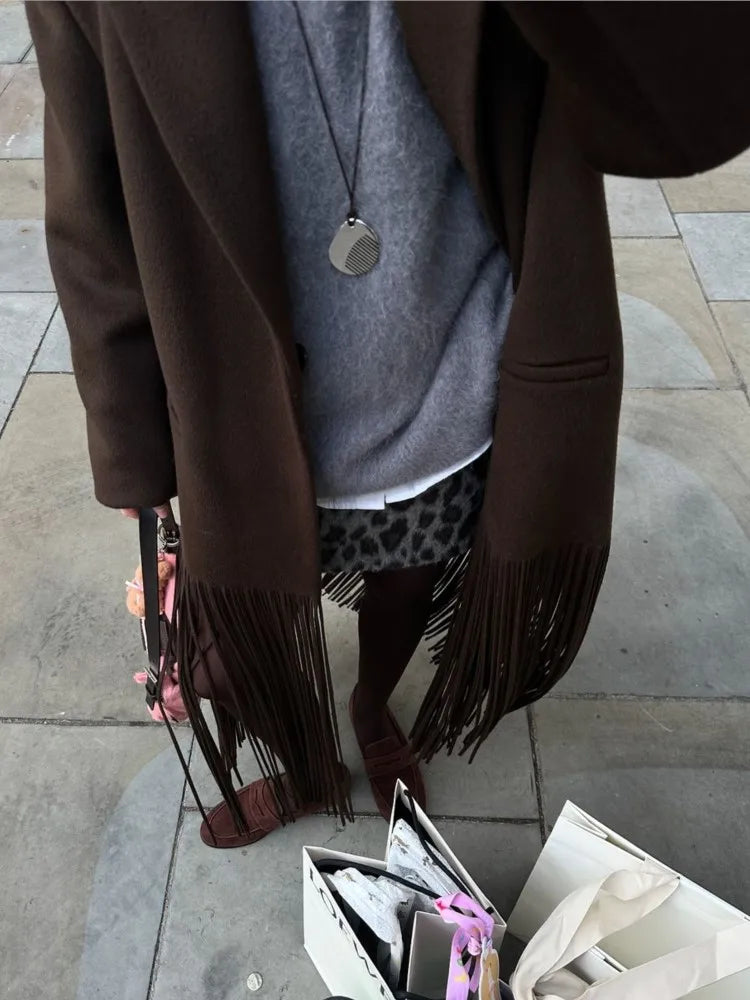 Person wearing a brown coat with fringes, gray sweater, and leopard print pants on a pavement.
