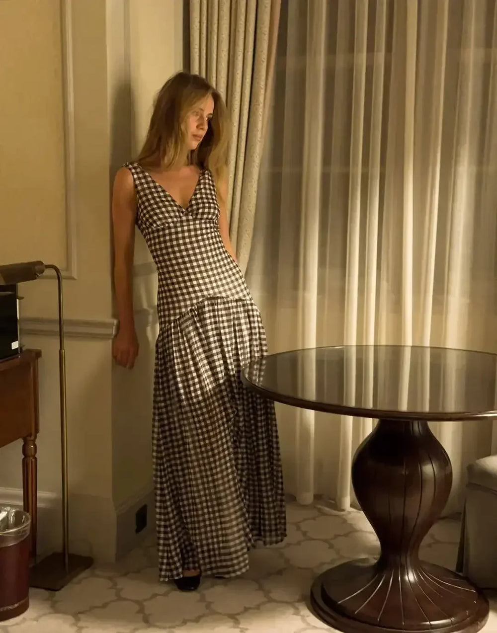 Woman in a checkered dress standing in a room with a table and curtains.