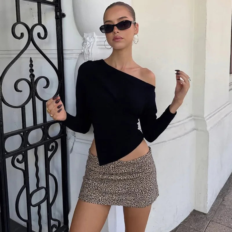 Woman wearing a black one-shoulder top and patterned skirt standing against a white wall.