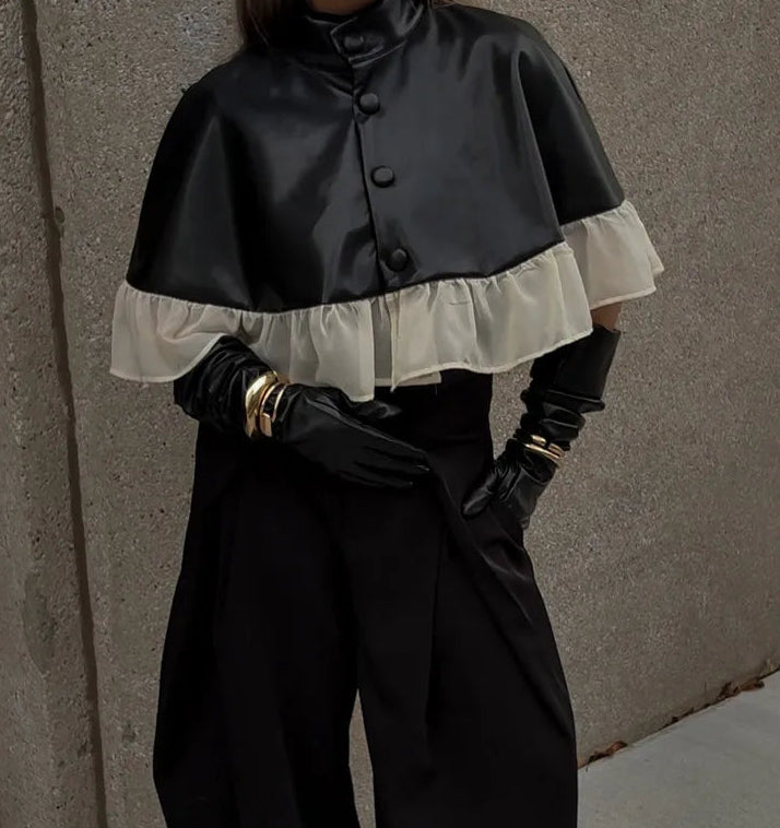 Person wearing a black cape with white ruffled trim against a gray wall.