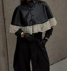 Person wearing a black cape with white ruffled trim against a gray wall.