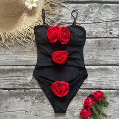 A black one-piece swimsuit with red rose embellishments laid flat on a rustic wooden surface, accompanied by a straw hat and flowers.