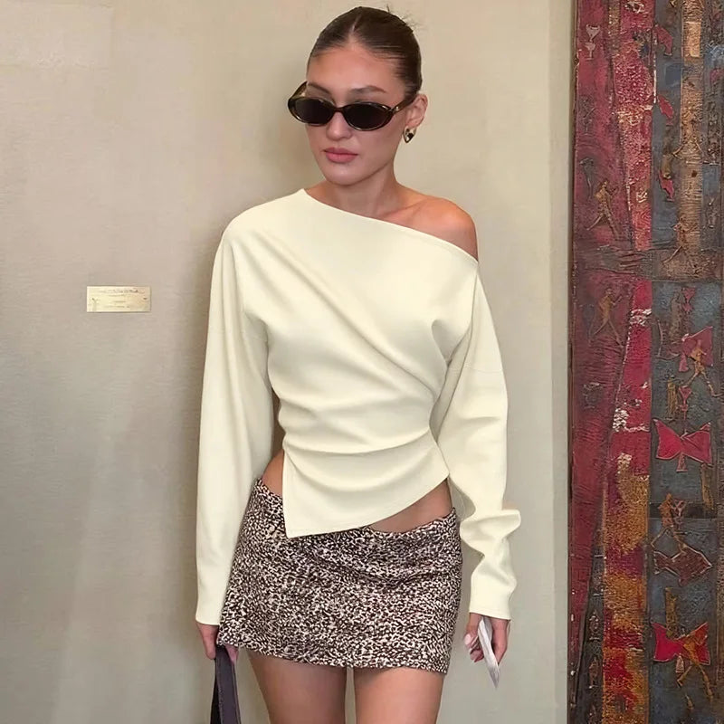 Woman wearing a white off-shoulder top and patterned skirt standing against a plain wall.