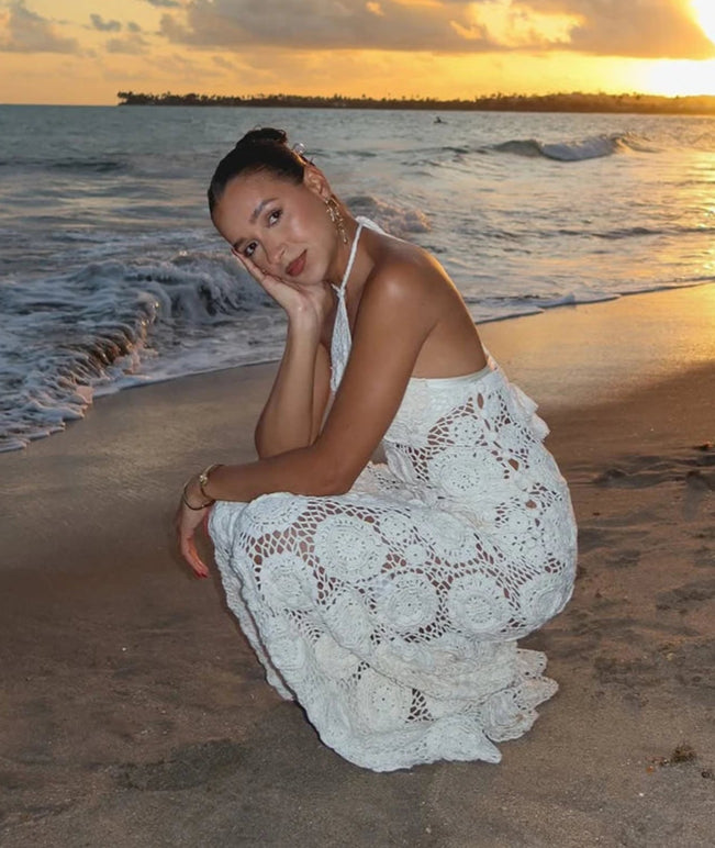 Woman in a white lace dress sitting on a beach at sunset.