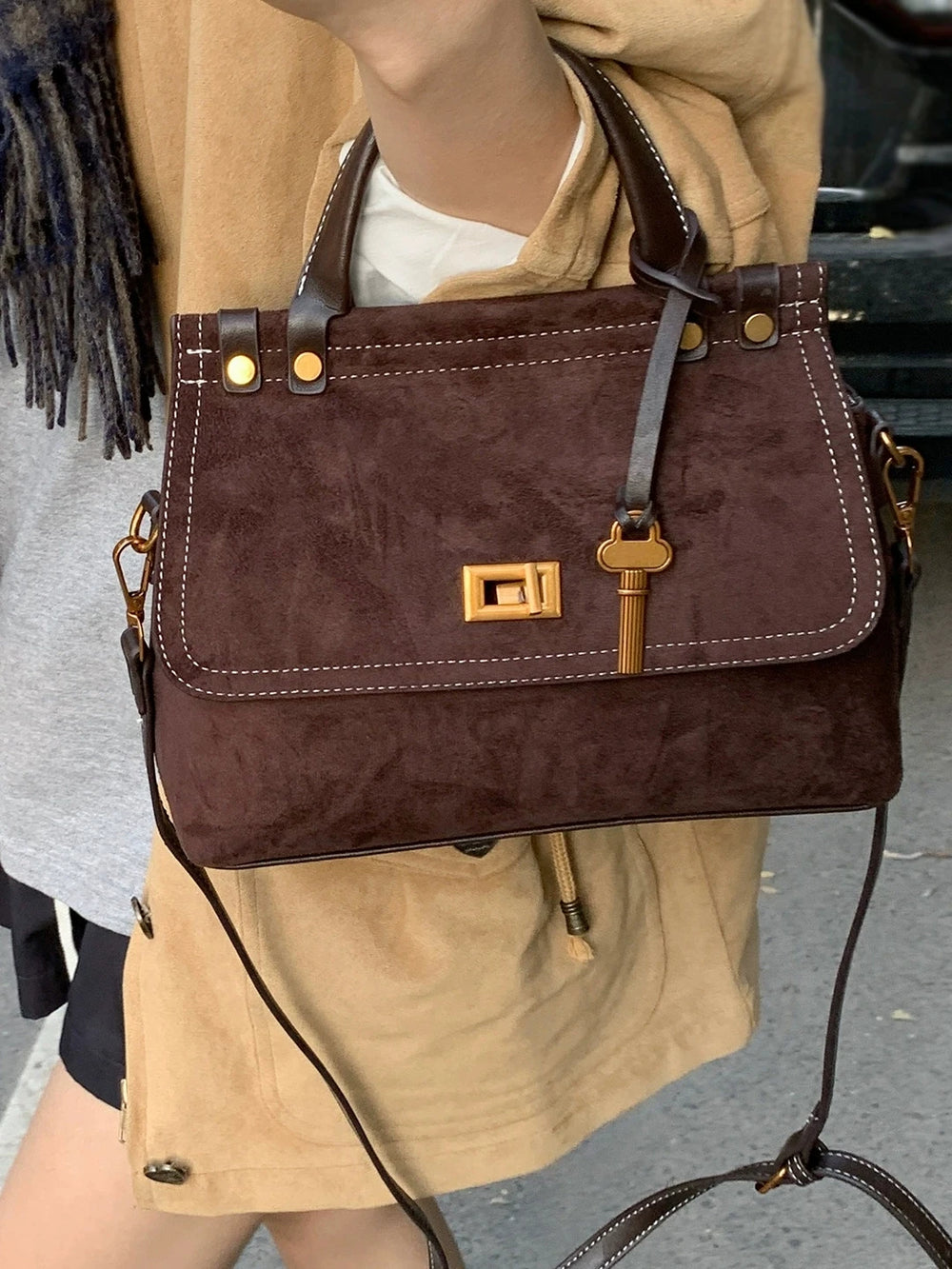 Brown handbag with gold accents held by a person wearing a beige jacket.
