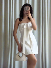 Woman wearing a white strapless dress with a blurred face, standing against a white curtain background with 'Trafza' branding.