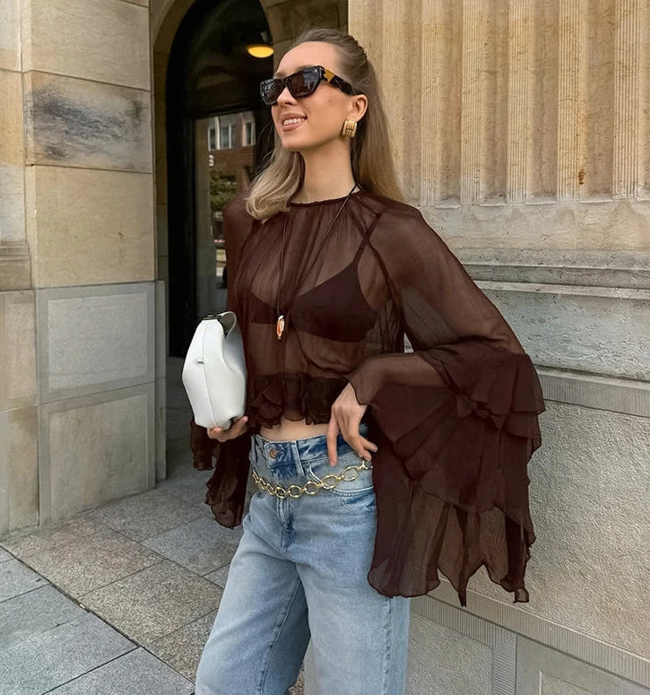 Woman wearing a sheer brown top and blue jeans standing in front of a stone building.