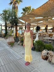 Woman in a yellow dress standing on a sandy path with palm trees and outdoor seating area in the background.