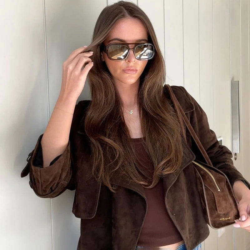 Woman wearing a brown jacket and sunglasses, holding a matching handbag against a neutral background.