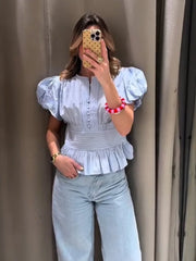 Woman taking a mirror selfie wearing a light blue top and jeans against a beige wall.