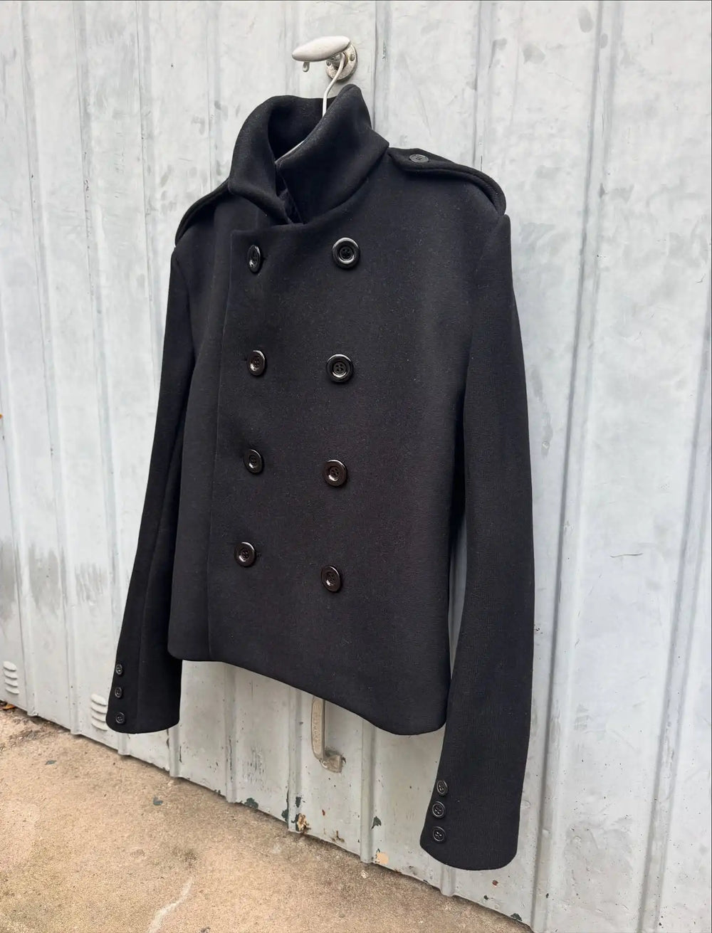 Black coat with double-breasted buttons hanging against a wooden wall.