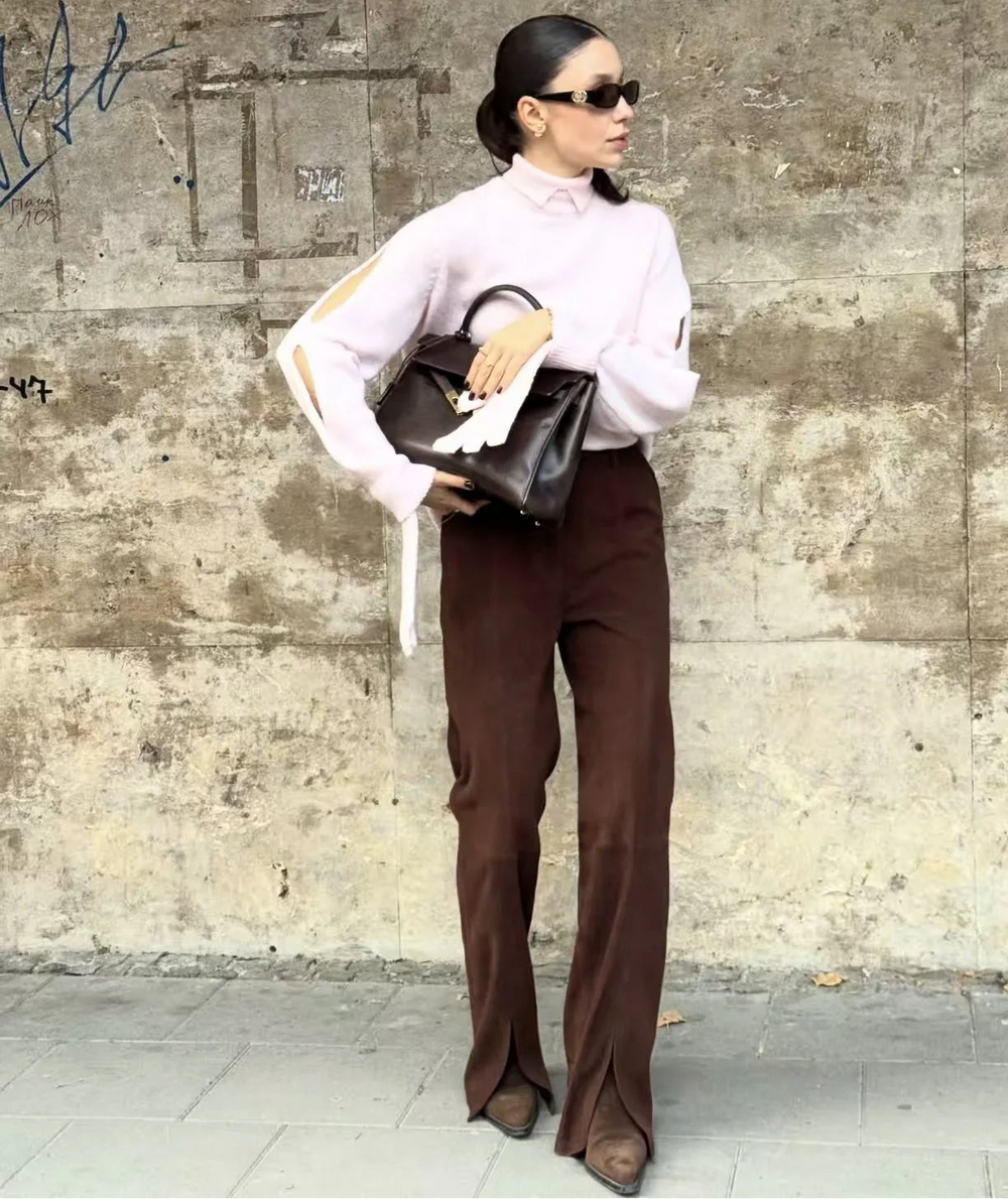 Person wearing a white blouse and brown pants against a textured wall.
