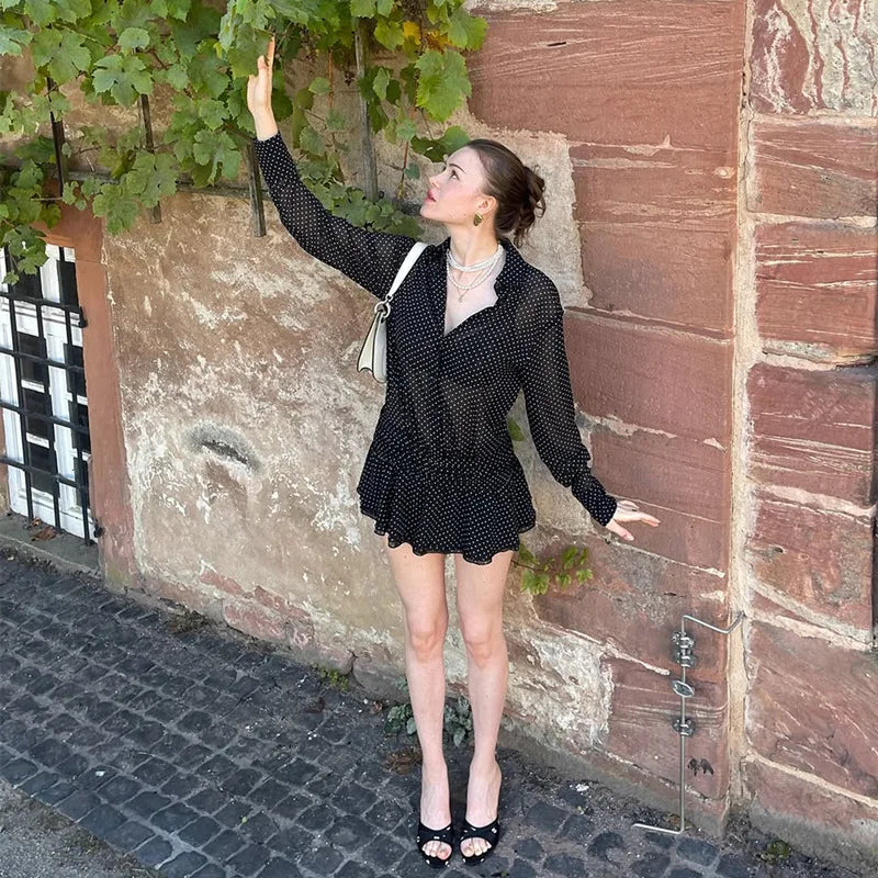 Woman in a black polka dot outfit standing against a textured wall with greenery.
