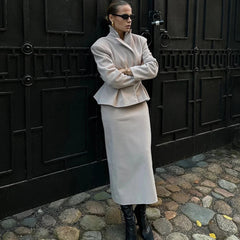 Woman in a beige coat and skirt standing against a dark, textured wall.
