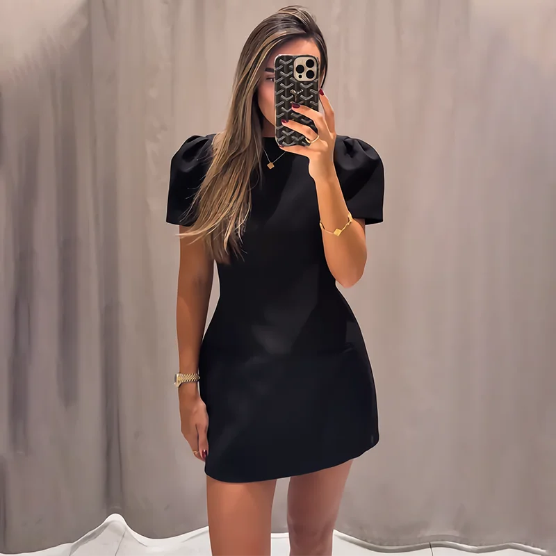 Woman taking a mirror selfie wearing a black dress against a plain background
