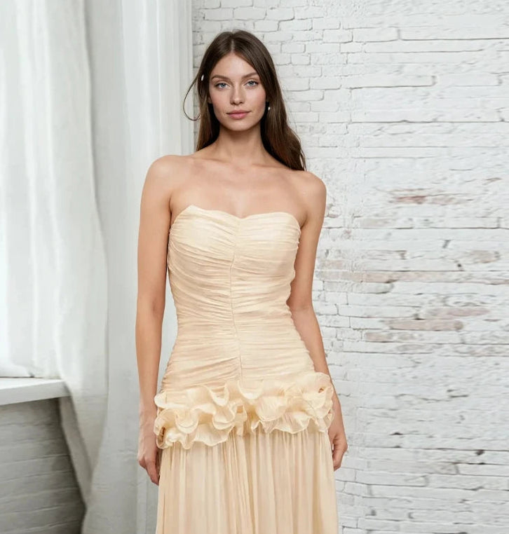 Woman wearing a strapless, ruched beige dress against a white brick wall.