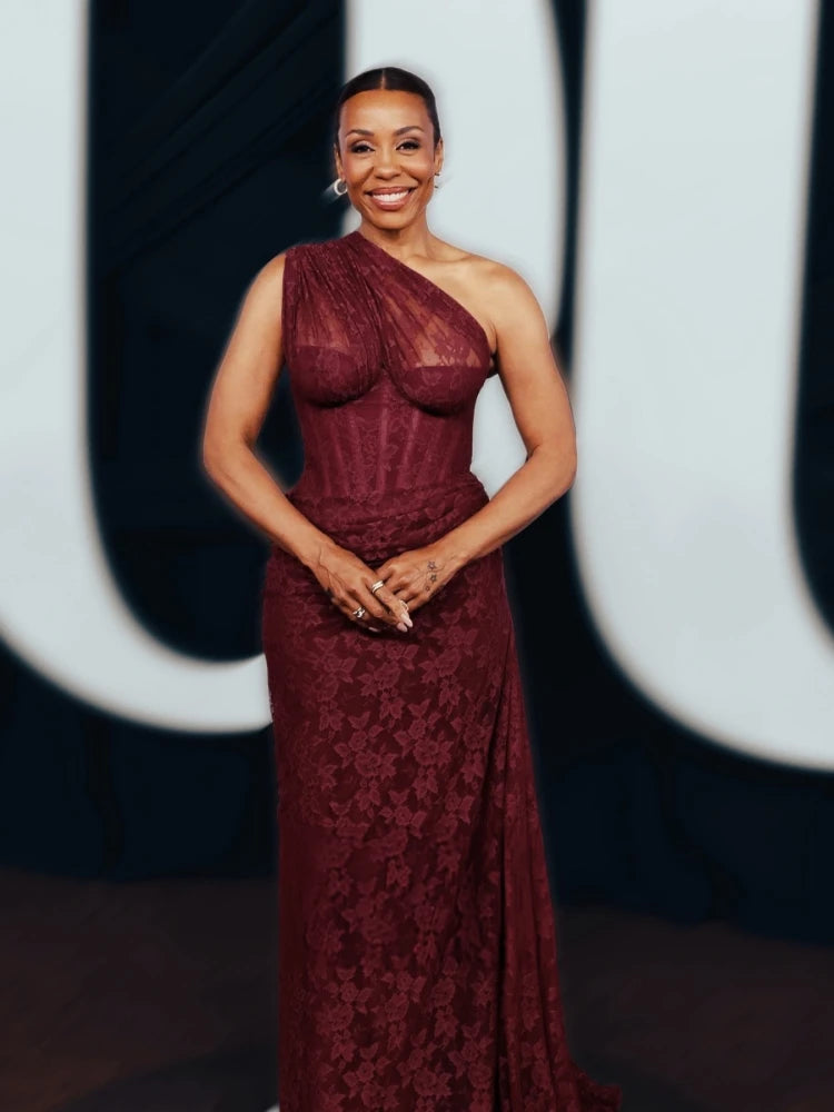 Woman in a burgundy lace dress standing against a blurred background with a visible logo.