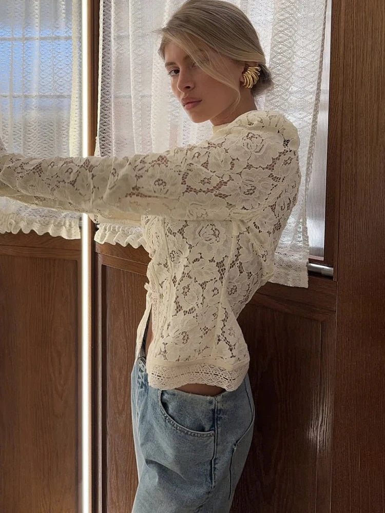 Woman wearing a white lace top and blue jeans standing in front of a window with lace curtains.