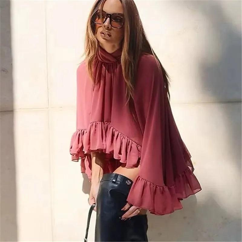 Woman wearing a pink blouse with ruffled sleeves and black pants against a neutral background
