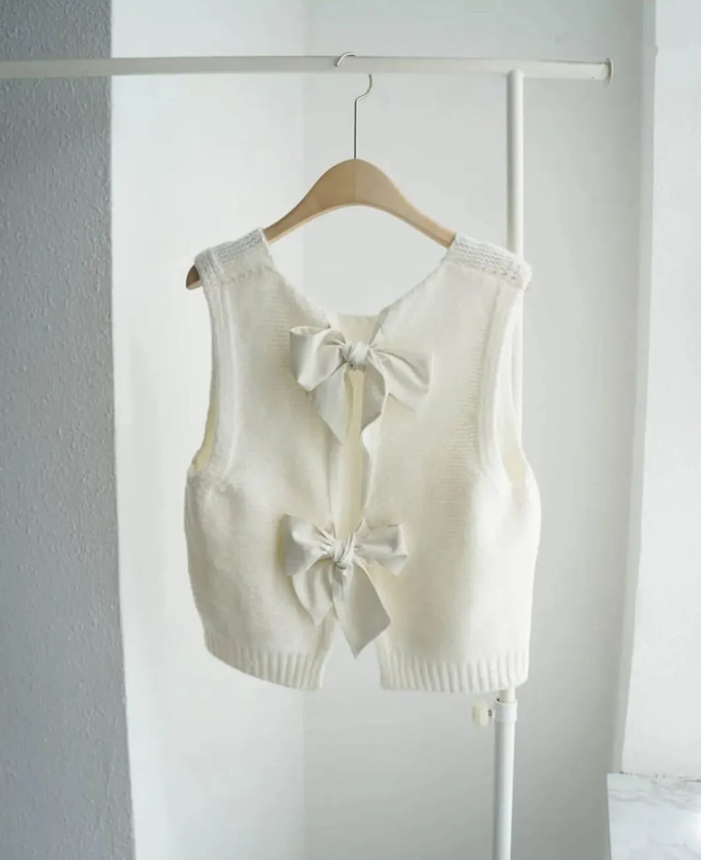 White sleeveless vest with bow details on a hanger against a white wall.