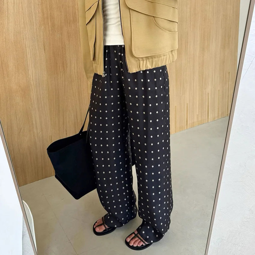 Person wearing a beige jacket, white shirt, black polka dot pants, and sandals, holding a black bag.