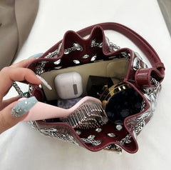 Hand holding a decorative bag with chain details, containing an Apple AirPods case and phone.