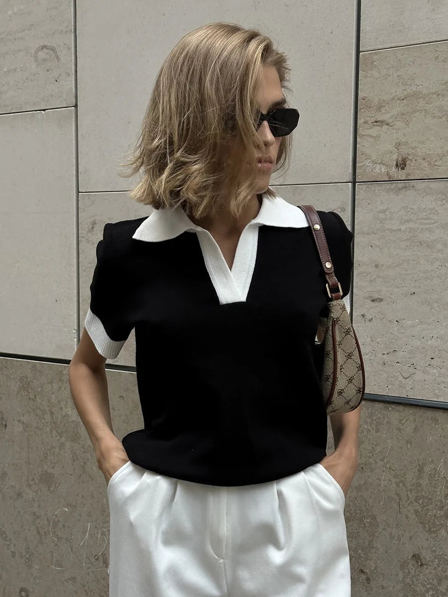 Woman wearing a black and white outfit with sunglasses and a handbag against a tiled wall.