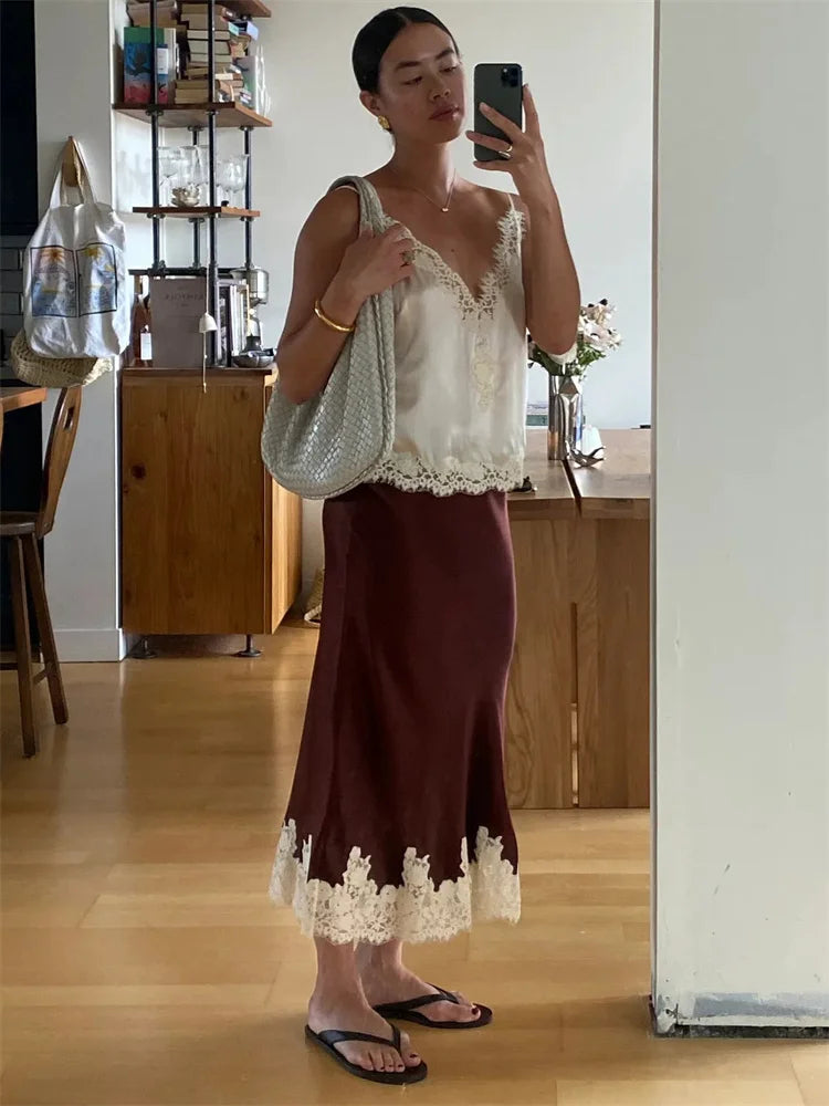 Woman taking a mirror selfie wearing a white lace top and brown skirt in a room.