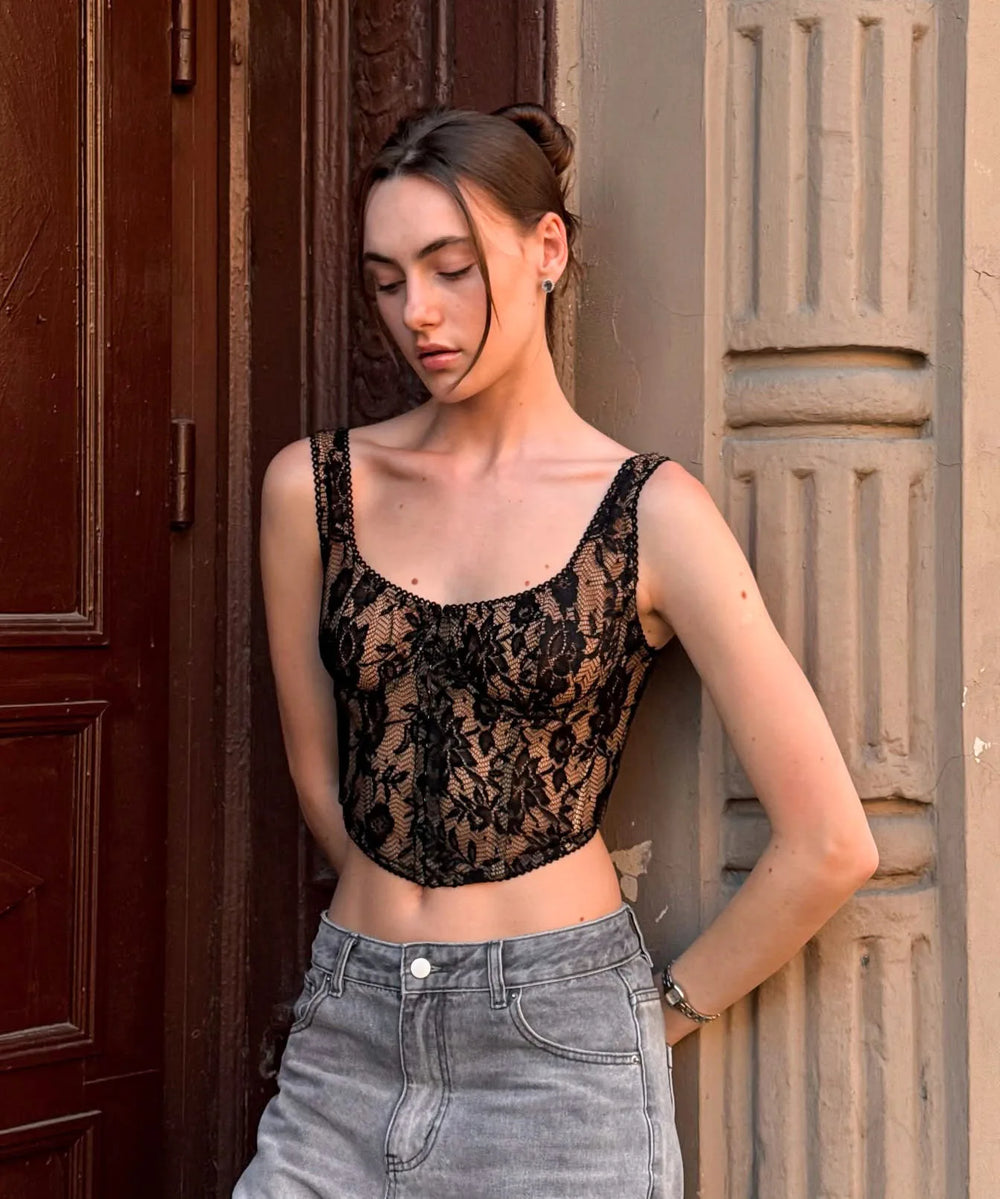 Woman wearing a black lace top and gray pants standing against a textured wall.