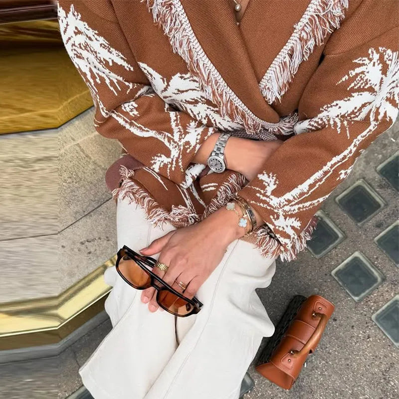 Person wearing a brown patterned coat with white fringes, holding sunglasses and a brown handbag on a street.