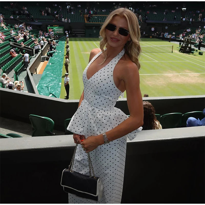 Woman in a white polka dot dress standing on a tennis court with spectators in the background