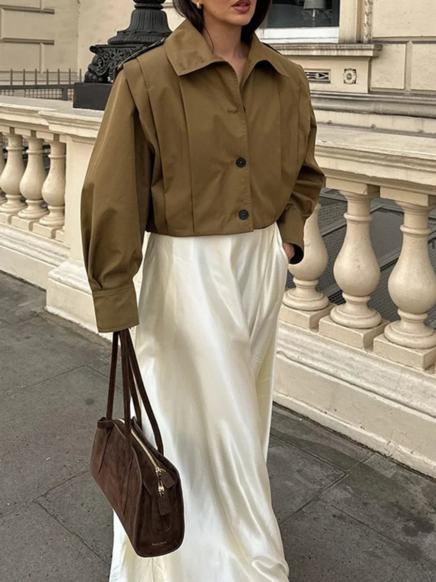 Person wearing a brown jacket and white skirt with a brown handbag, standing in front of classical architecture.