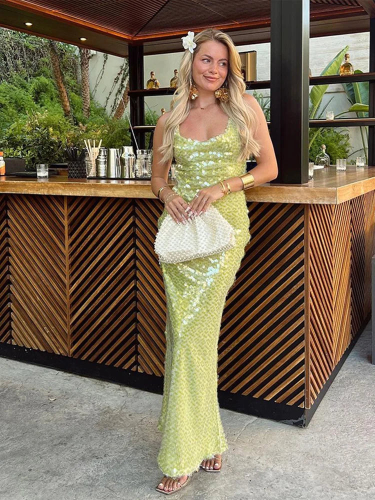 Woman in a green sequin dress holding a white clutch in front of a wooden bar.